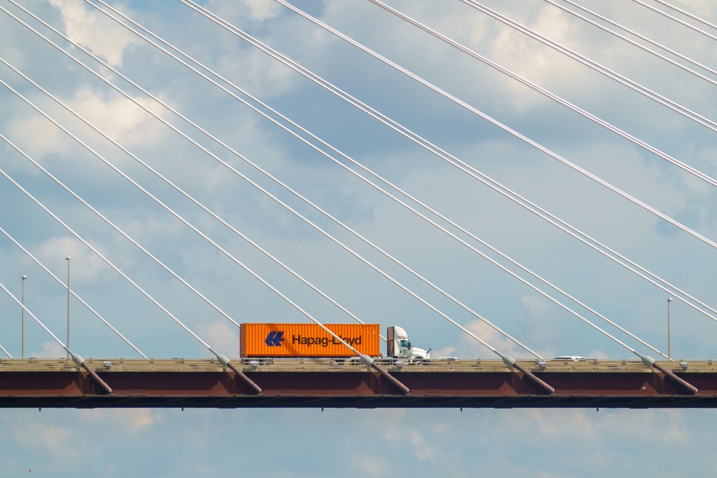 Orange Hapag-Lloyd shipping container truck crossing a cable-stayed bridge beneath diagonal suspension cables and blue sky.